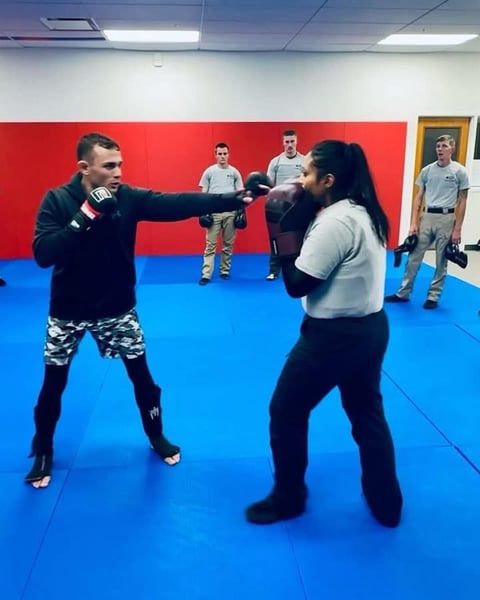 Two martial artists sparring in a boxing ring during a training class with instructors and students watching in the background