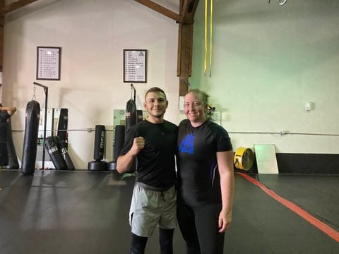 Two men posing together in a gym with equipment and training materials visible on the walls in the background