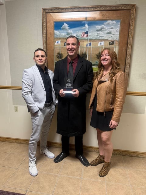 Three people pose together indoors, with a man in the center holding an award