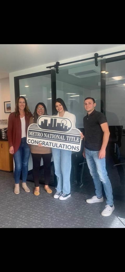 Four people holding a Metro National Title congratulations sign in an office with glass doors and warm lighting