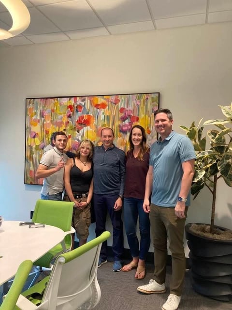 Five people standing together in an office space with a colorful flower painting, green chair, and potted plant visible